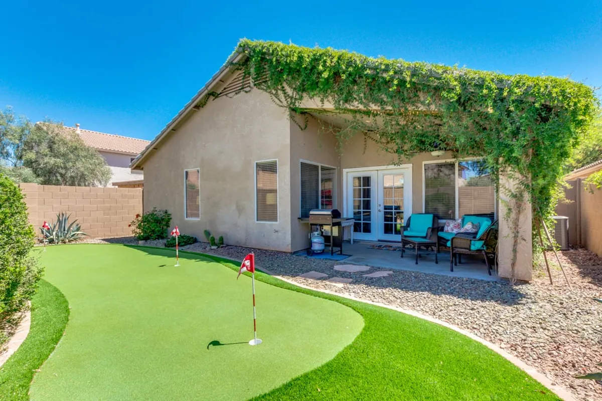 A spacious backyard featuring a well-maintained artificial putting green, outdoor seating for dining and lounging, and lush greenery framing the area. The setup invites both recreational golf practice and relaxation, capturing the essence of indoor-outdoor living.