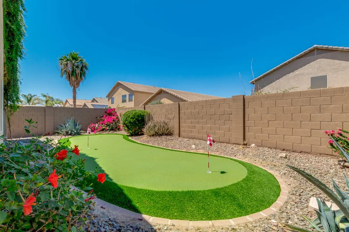 A well-manicured backyard featuring an artificial putting green, vibrant flowers, a palm tree, and privacy walls, offering ample space for outdoor activities like golf practice, relaxing, or hosting small gatherings.