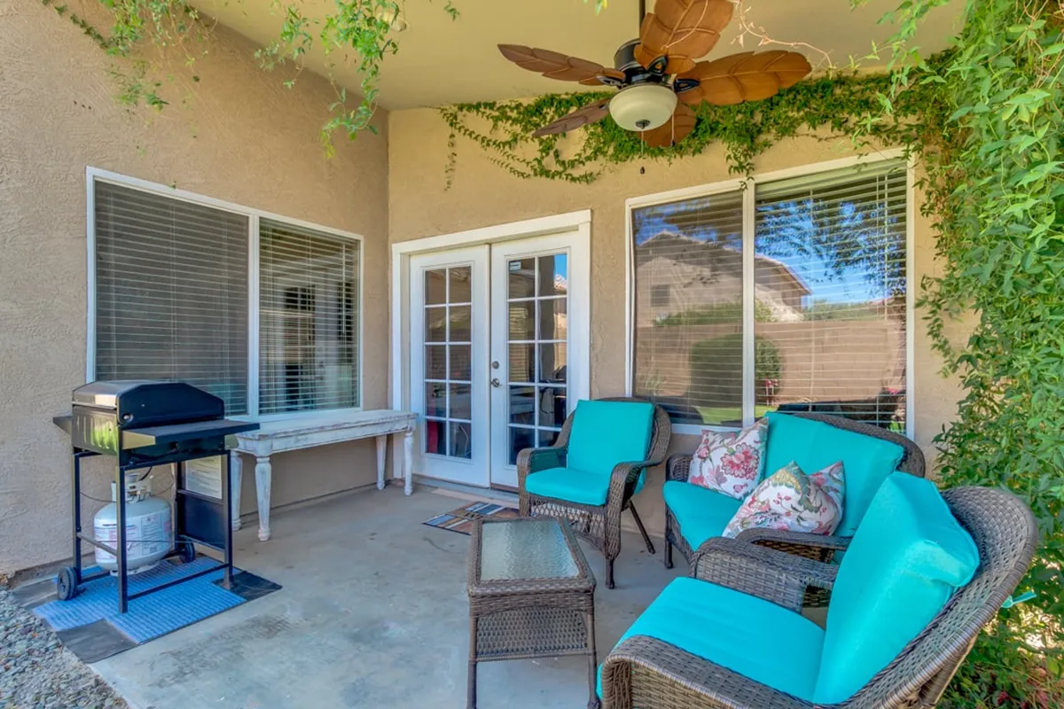 A cozy outdoor room setup with turquoise chairs around a glass table, a grill for outdoor cooking, and lush greenery. The double doors suggest easy transition from the indoors, reinforcing the sense of flow between interior and exterior spaces.