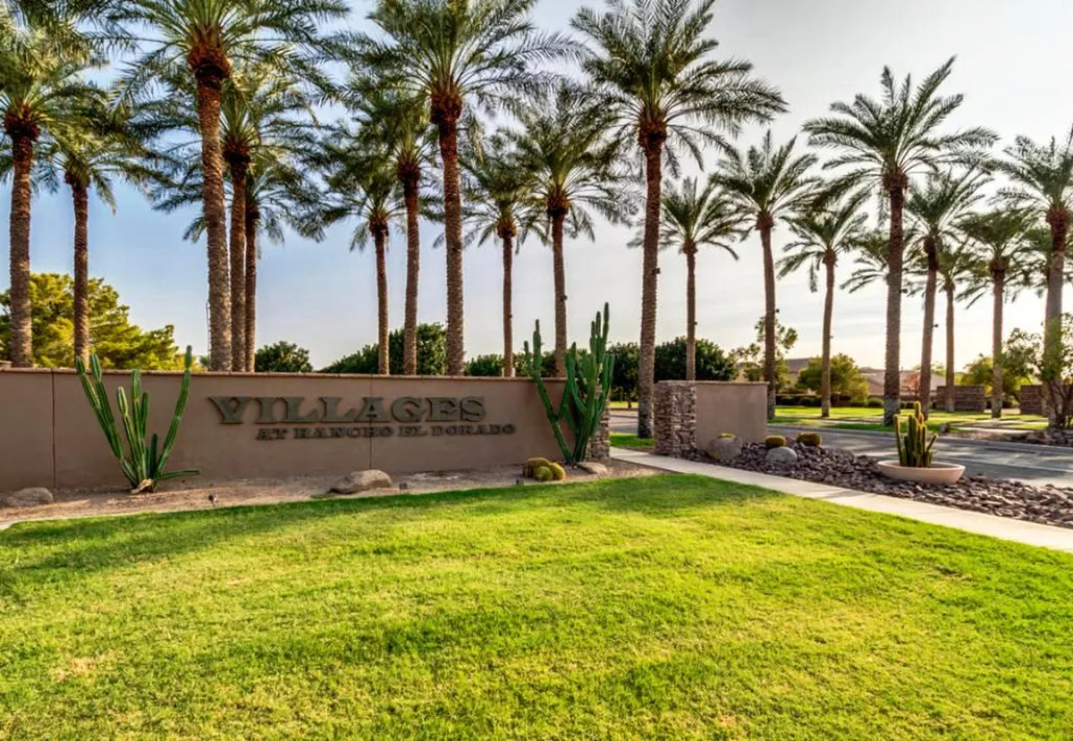 A welcoming exterior view of the property, featuring clear signage, swaying palm trees, a manicured green lawn, and desert cacti. The scene exudes a sense of arrival and hints at the vibrant lifestyle within the community.