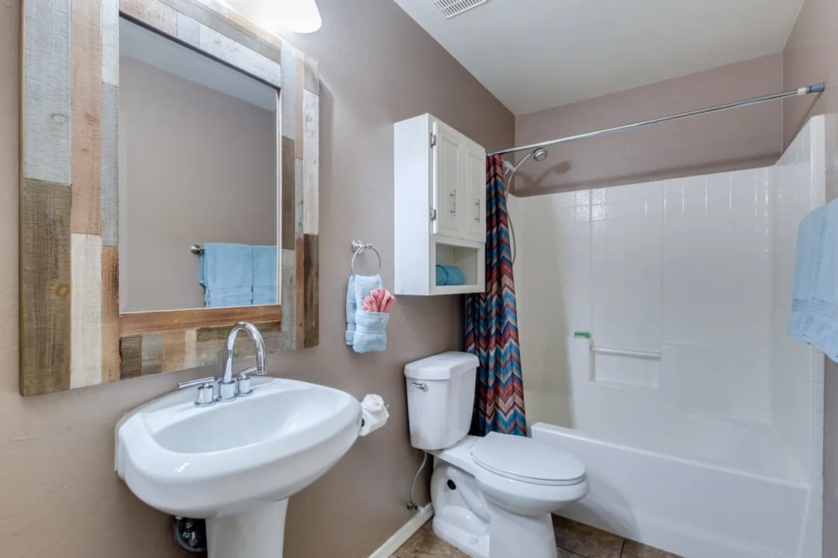 A bright shared bathroom featuring a decorative shower curtain, framed mirror, modern sink, and toilet—offering a practical and inviting space for guests.