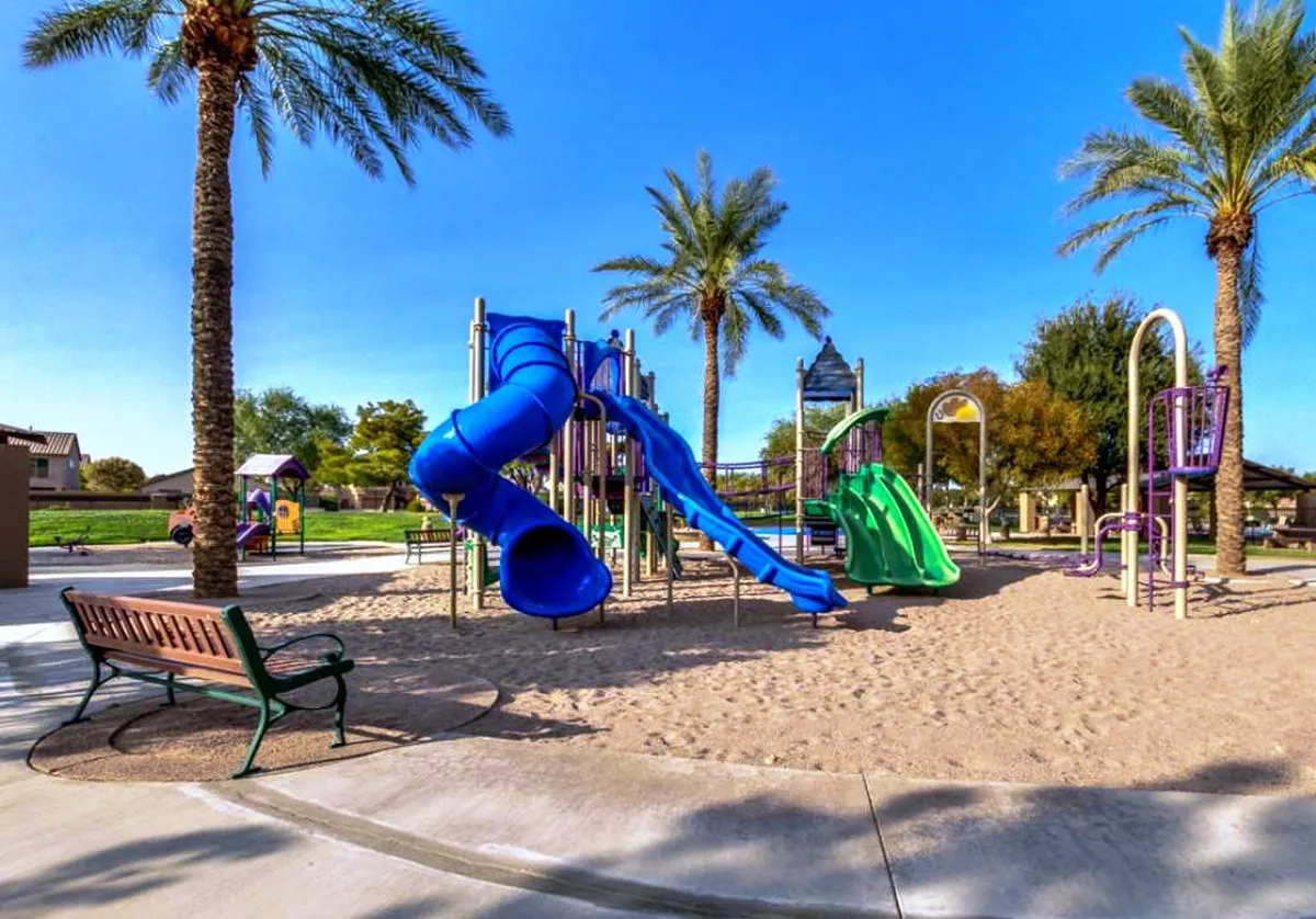 A vibrant playground featuring colorful equipment surrounded by palm trees and benches, with plenty of space for children to play and families to gather. The cheerful atmosphere is enhanced by bright sunlight and greenery.