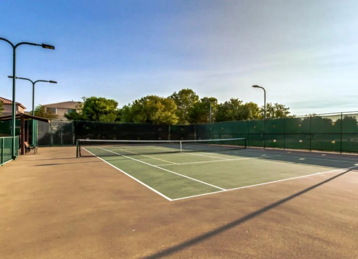 The image showcases a vibrant tennis court set amid mature trees and well-kept surroundings. Lighting poles indicate the space can be enjoyed both day and evening, fostering a dynamic, energetic atmosphere for recreation and fitness.