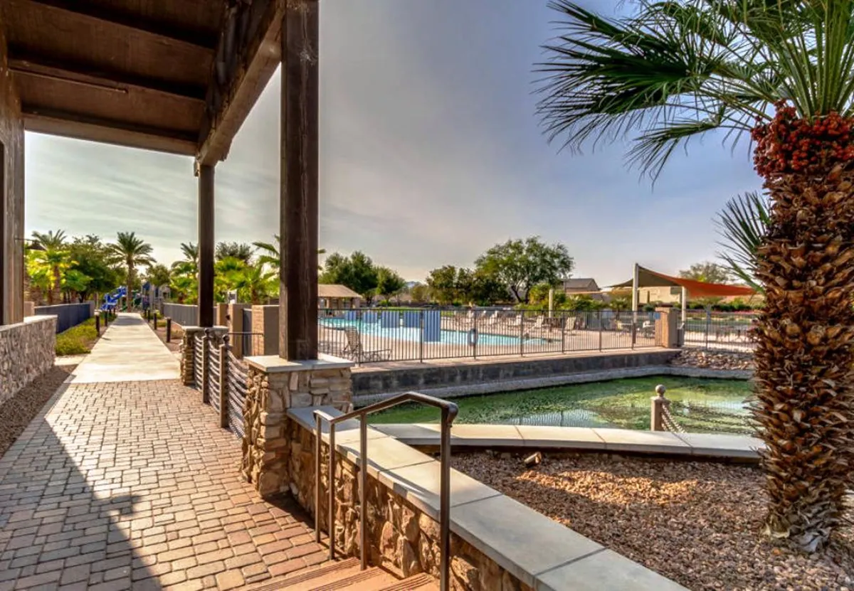 A serene and inviting pool area featuring a paved walkway, palm trees, and a tranquil water feature, all surrounded by fencing for privacy and safety. The scene hints at a laid-back, resort-style lifestyle.