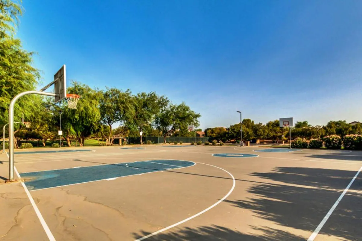 An outdoor basketball court with multiple hoops and a well-maintained surface, bordered by lush trees. The space is clearly designed for active recreation and social meetups.