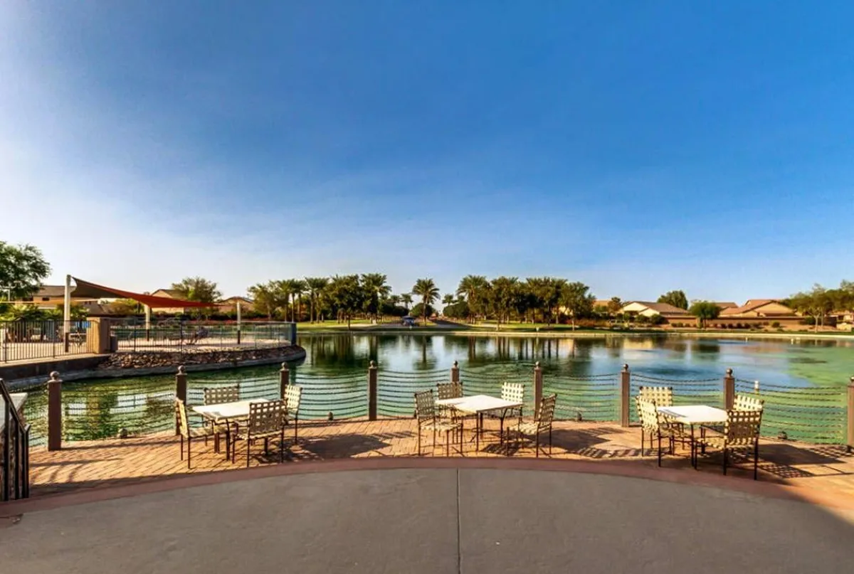 This exterior scene features a serene lake view with outdoor seating under a sunshade, framed by lush trees and lawns. The image captures the tranquil ambiance of the resort community and opportunities for relaxation or social gatherings by the water.