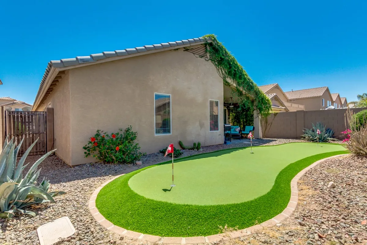 A backyard view centered on a putting green bordered by lush plants and decorative greenery. The layout invites guests to practice golf, relax, or host small, casual gatherings in a tranquil outdoor setting.
